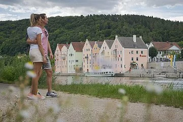 Ein Pärchen in sommerlicher Kleidung steht am Main-Donau-Kanal und halten sich im Arm. Sie schauen aufs Flussufer gegenüber, dort ist die Riedenburger Promenade zu sehen. Dahinter ragen die Hänge mit ihrem dunkelgrünen Kleid auf.